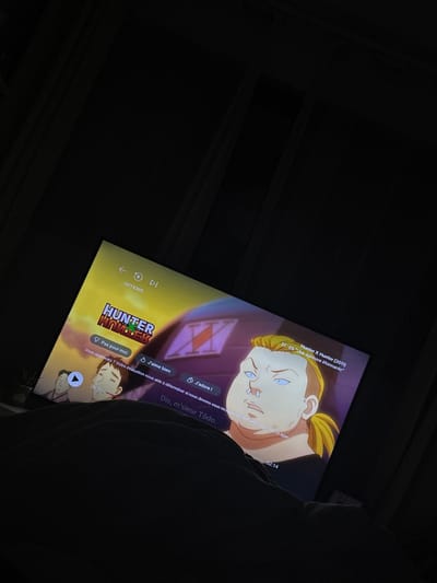 The image displays an indoor scene in Villecresnes, France, characterized by low light conditions. A television screen, positioned at an upward angle from the viewer's perspective, dominates the lower half of the frame. The screen is displaying content from the anime series "Hunter x Hunter (2011)," specifically Season 1, Episode 6, titled "Une épreuve étonnante" (A Surprising Trial).

On-screen, the title "HUNTER x HUNTER" is visible on the left. User interface elements in French include "OPTIONS," playback controls (back, play/pause, forward), and interactive prompts such as "Pas pour moi," "J'aime bien," and "J'adore!" A feedback message states, "Vous appréciez ? Votre évaluation nous aide à déterminer si nous devons vous recommander ce type." A subtitle at the bottom reads, "Dis, m'sieur Todo." Two animated characters are depicted: a male character with blond hair, a ponytail, blue eyes, a nose ring, and a robust build occupies the right side of the screen, while a smaller, brown-haired male character is partially visible on the left. Timestamps "20:25" and "02:14" are also present. The bottom edge of the screen is partially obscured by a dark, indeterminate soft object, likely bedding, suggesting a person is watching from a recumbent position. The upper portion of the image is dark, revealing faint vertical patterns consistent with window blinds or curtains.
