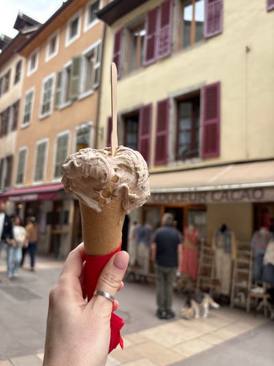In Annecy, France, a person's hand holds a cone of chocolate gelato topped with swirls of lighter cream and dark chocolate chips. The gelato is served in a waffle cone, which is wrapped in a bright red paper holder. A silver ring is worn on the index finger. In the background, a narrow street is lined with buildings featuring peach and pale yellow facades, adorned with purple shutters. A sign on one building reads "COULEUR CACAO". Several people are visible in the background, some blurred and walking, while mannequins display clothing outside a shop. A small dog is also present on the cobblestone street.