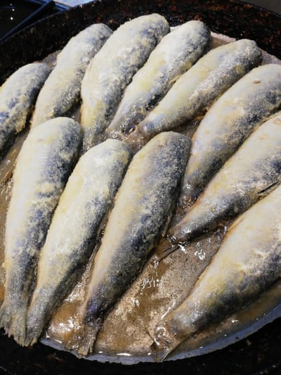 The image shows a close-up, overhead view of several small, whole fish, likely sardines or herring, arranged in a circular pan. The fish are coated in a light-colored powder, possibly flour or cornmeal, and are frying in oil, evident from the bubbling liquid surrounding them. The pan appears to be dark, possibly cast iron, with a textured surface and some dark residue on the edges, suggesting it has been used for cooking. The fish are lined up in rows, with their heads generally pointing towards the top left and their tails towards the bottom right. The lighting is bright, highlighting the glistening oil and the slightly opaque texture of the fish. There are no people or any other objects visible in the image, and no specific location within Новапагосцкі сельскі Савет, Belarus can be identified.