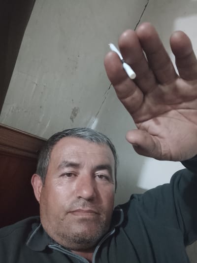 The image depicts a middle-aged man with short, graying hair, holding a white cigarette or similarly shaped object in his outstretched hand. His hand is in the foreground, partially obscuring the background. The man is wearing a dark-colored shirt with a light-colored collar. The background consists of a worn-looking wall, possibly painted white, with visible cracks. Based on the prompt, the location is Sabir Rakhimov, Uzbekistan, but no specific landmark or detail visible in the image confirms this location.