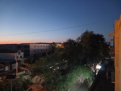 This media file captures an urban scene at dusk or dawn, with a clear sky transitioning from a deep blue to a faint orange near the horizon. The view is from an elevated position, likely a balcony or rooftop, looking out over a street and buildings.
In the foreground, a textured rope is visible, adding a tactile element to the scene. Further out, a residential or commercial building with multiple windows stretches across the mid-ground. Adjacent to it, a smaller, distinct white building with a domed roof and arched windows stands out. Trees with dense foliage are scattered throughout the scene, some illuminated by unseen lights, creating a dappled effect. A power line stretches horizontally across the sky, a common urban element.
The overall setting suggests a town or city, possibly Qorowul, Uzbekistan, given the provided location context. The lighting indicates either early morning or late evening, with artificial lights from within buildings and possibly streetlights beginning to glow. The atmosphere is calm and quiet, typical of transitional periods of the day. No people are clearly visible, focusing the attention on the architecture and natural elements. The scene is devoid of any apparent event or activity, presenting a still, observational view of the urban landscape.