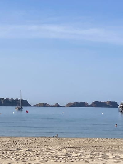 The image depicts a serene coastal scene in Peguera, Spain, likely captured from a sandy beach. The foreground is dominated by a textured, sun-drenched sand dune area, with footprints visible, suggesting recent activity. Beyond the beach, a calm, blue sea stretches towards a distant horizon.
In the water, a white sailboat with two masts is anchored, its sails furled. A red buoy is positioned near the sailboat. Several other buoys, appearing yellow, are sparsely scattered across the water's surface. In the mid-ground, a person is swimming, their head visible above the water, and another individual appears to be on the deck of a white vessel to the far right of the image.
The background features a chain of rocky, uninhabited islands with scrubby vegetation, under a clear, pale blue sky. Buildings are visible on the leftmost part of the island chain, suggesting the presence of the Peguera coastline. The overall atmosphere is tranquil and idyllic, characteristic of a Mediterranean seaside resort.