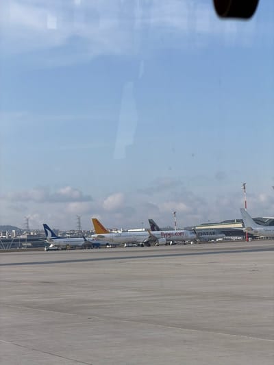 The image displays an airport tarmac scene under a partly cloudy blue sky, captured through a reflective window. In the midground, several commercial passenger aircraft are parked. From left to right, a white aircraft with a blue vertical stabilizer, consistent with a Turkish Airlines or AnadoluJet livery, is visible with ground support equipment near its landing gear. Next to it, two white Pegasus Airlines aircraft are present; one features 'PEGASUS' on its yellow tail, and the other has 'flypgs.com' written on its fuselage. Behind the latter, a white aircraft with a maroon tail emblem and 'QATAR' on its fuselage, identifying it as a Qatar Airways plane, is partially obscured. The concrete tarmac extends into the foreground. In the background, modern airport terminal buildings, jet bridges, and various ground support vehicles are visible. Beyond the airport infrastructure, a distant urban landscape with multiple buildings and prominent lattice-style electricity transmission towers extends towards the horizon. No individuals are discernible. The scene is consistent with operations at Sabiha Gökçen International Airport (SAW), located near Tuzla, Turkey.