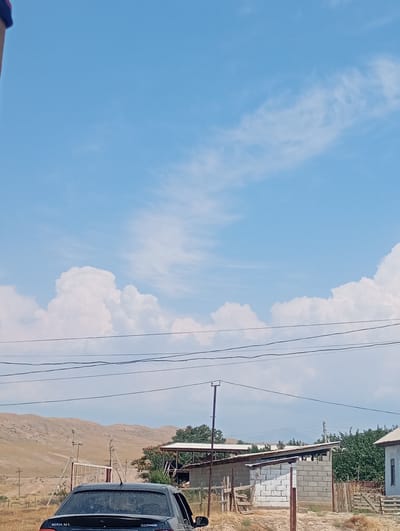 The image captures a scene in Koçkor-Ata, Kyrgyzstan, under a bright blue sky with scattered white clouds. In the foreground, a dark-colored sedan, possibly a Daewoo Nexia, is parked. The car's rear is visible, with a license plate that reads "NEXIA 1.6 L". Behind the car, a rural landscape unfolds. A rough, unpaved road leads towards a gate structure made of metal poles and fencing. Further back, rolling, dry hills dominate the horizon. To the right, there are some low-lying buildings constructed with concrete blocks, and dense green trees provide a backdrop. Power lines are strung across the midground of the image. The overall impression is one of a sunny day in a semi-arid, rural environment.