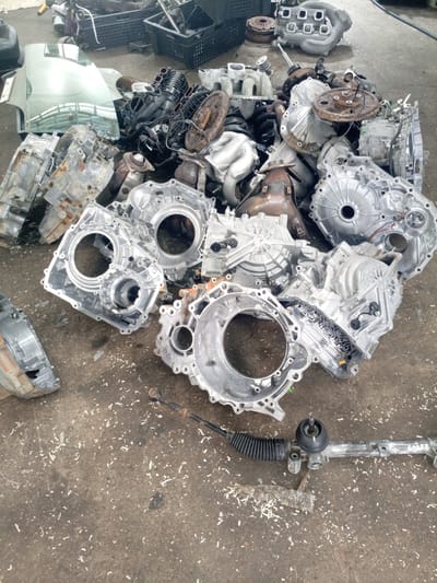 The image depicts a junkyard or auto salvage yard in Oktyabr'skii, Russia. The ground is concrete and littered with various automotive parts, primarily transmissions and engine components, which are scattered in a haphazard pile. Some of these parts are made of aluminum and appear to be somewhat aged and dirty. In the background, there are black plastic crates and other scrap metal. In the foreground, a steering rack with a protective bellows is visible. There are no people or animals present in the image. The overall impression is one of disarray and the accumulation of discarded vehicle parts.