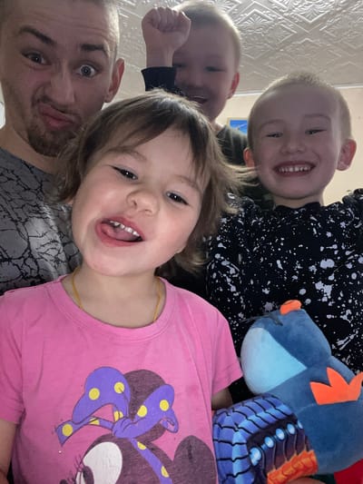 A selfie photograph, possibly taken in a residential setting (given the wall texture visible), features a man and three children. The man is positioned in the lower-left corner of the frame, displaying a surprised expression. One child is in a pink Minnie Mouse shirt. Another child is wearing a black and white splatter-patterned shirt. A blue stuffed animal is also visible. The image provides insufficient detail to confirm a location in the city of Bely Klyuch, Russia. The photo appears to capture a casual, familial moment.