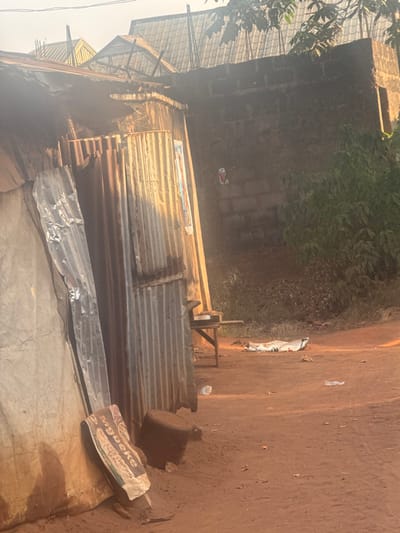 A corrugated metal structure sits beneath a clay-like wall. Nearby, a drum and labeled paper rests on a cloth-covered surface. A wooden mechanism stands beside the structure, on the dirt surface. The area is partly shaded by foliage above, with a building visible in the background. The atmosphere suggests a modest, possibly rural setting.