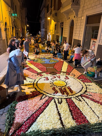 In a narrow, cobblestone street in Tarquinia, Italy, an evening scene depicts numerous individuals constructing a large, elaborate floral carpet. The street is flanked by multi-story buildings, illuminated by warm artificial light from street lamps and shop windows, against a dark night sky.

The central subject is a multi-segment floral carpet, meticulously assembled from flower petals of various colors, covering a significant portion of the street's width and extending into the background. The visible portion of the carpet features a large circular design with a yellow center, radiating red and white segments, and dark outlining. Within the yellow center, red letters are arranged, appearing to spell "SHI" or "IHS." Beyond this, the carpet continues with geometric patterns in pink, red, white, and brown petals, and further back, a blue and green design resembling a globe. Bundles of green foliage and stems line the edges of the carpet on the ground.

Approximately two dozen people are visible, actively participating or observing. In the foreground, an older woman dressed in a light blue habit and white coif (a nun) kneels, carefully placing white flower petals onto the carpet with her right hand. She wears light-colored boots. To her right, in the midground, a man in a patterned short-sleeved shirt holds a cardboard box labeled "cucinArtig" and pours white petals into a green plastic bucket on the ground. Other participants, casually dressed, are observed kneeling, bending, or standing across the carpet, many with hands near the petals, suggesting active involvement in its creation. Some hold small containers or refer to a design plan held by a man in the background.

The surrounding environment includes architectural details such as windows, balconies, and entryways on the buildings. On the left building, a green digital display indicates "21 °C." On the right building, a vertical sign reads "ZANZARIERE E PORTE" (Mosquito nets and doors), with a smaller advertisement below it. Various cardboard boxes, some filled with petals, and plastic buckets are scattered along the edges of the carpet, serving as containers for the materials.
