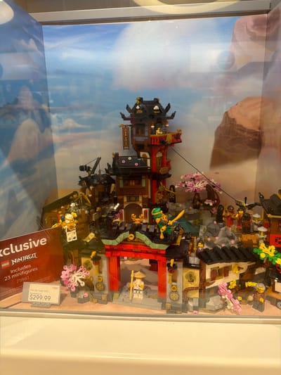 The image shows a LEGO Ninjago "The Old Town 15th Anniversary" set priced at $299.99, displayed in a store window. The set features a detailed replica of an ancient Japanese town with multiple buildings, including a multi-story pagoda-like structure, a traditional gate, and smaller houses. Several LEGO minifigures are integrated into the scene, some appearing to be in combat poses, with others standing guard or observing. Cherry blossom trees, depicted with pink LEGO pieces, add a decorative element to the landscape. A price tag and a product label indicating "Exclusive," "LEGO Ninjago Legacy," and "23 minifigures" are visible in the foreground. The backdrop of the display appears to be a printed scene of a cloudy sky and rocky terrain, suggesting an outdoor, historical setting. The display is framed by a grey border at the top and a white surface at the bottom, indicative of a retail environment. This display is likely located within the city of Mission Viejo, United States of America, as indicated by the user's prompt.