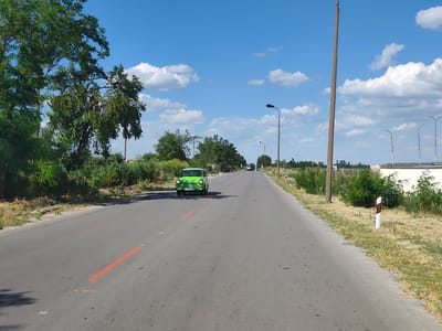A bright green, vintage car is driving down a paved road. The car is centrally located in the frame, driving towards the viewer on the road. The road has orange markings on it. On either side of the road are green trees and bushes, with dry, yellow grass. In the background, there are a few more trees and what appears to be a white building with a fence. The sky is blue with scattered white clouds. There are several utility poles visible along the right side of the road, some with streetlights. One utility pole on the right is very tall and rustic. There are also road markers on the right side of the road with red and white stripes. The scene appears to be set in a rural or suburban area. The time of day appears to be daytime, likely mid-morning or afternoon, given the bright sunlight and shadows. The weather appears to be clear and sunny. The license plate on the car reads "CH 7624 MS".