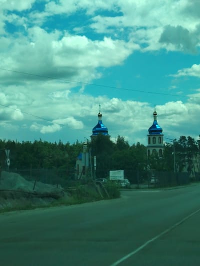 The image shows a view from a moving vehicle on a paved road. In the background, there are two domed buildings, likely churches, with blue roofs and golden spires, surrounded by lush green trees. A smaller white building with a blue roof is also visible. The sky is filled with dramatic clouds against a bright blue backdrop. On the left side of the road, there is a pile of sand and a black metal fence. Several road signs are present, including a white circular sign with a red border and a black symbol. The overall scene suggests a rural or semi-rural setting in Kapitanivka, Ukraine, during daylight hours.