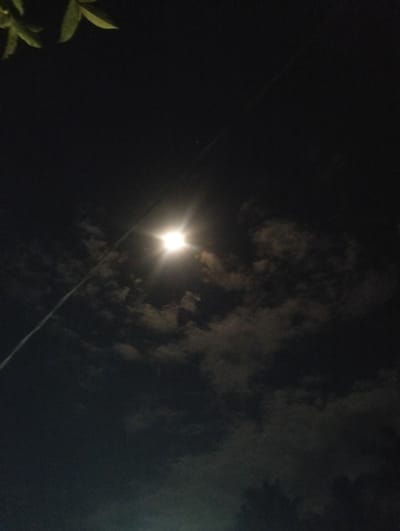 The image depicts a night scene in Peuneulet Baroh, Indonesia. The sky is dark and cloudy, with a bright light source, likely the moon, shining through the clouds. The light creates a halo effect. A power line is visible diagonally across the sky. A few leaves are visible in the upper left corner, and blurry trees are visible at the bottom of the frame.