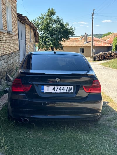 The image shows the rear of a black BMW sedan parked on a grassy area next to a brick building. The license plate reads "T 4744 AM" with a "BG" indicator. In the background, there are houses with tiled roofs, a wooden utility pole, and a stack of firewood. The sky is blue with some clouds. The location is identified as Konop, Bulgaria. There are no people visible in the image.