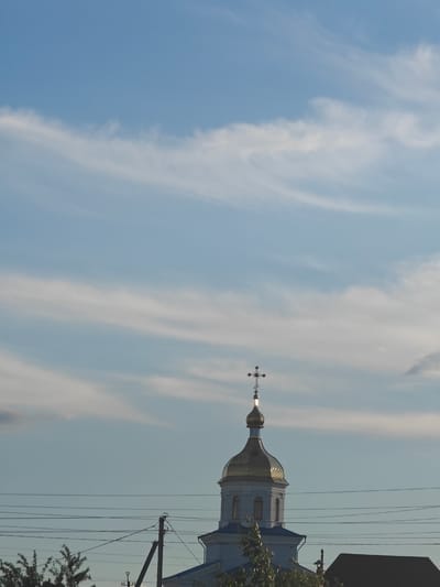 The image shows a light blue sky with wispy white clouds. In the lower portion of the image, a golden-domed church with a cross on top is visible. The church has light blue roof elements and white walls. In front of the church, there are power lines and a utility pole. Trees are also visible in the foreground. The scene appears to be set in a rural or semi-rural area of Kotelva, Ukraine, likely on a clear day with some cloud cover.