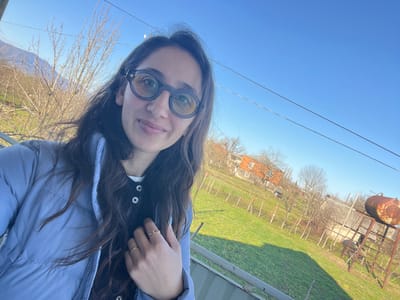 The image displays a female individual taking a selfie outdoors in Zeda Sakara, Georgia, under a clear, bright blue sky. The subject, positioned in the upper left quadrant, has long, dark, wavy hair and wears dark-rimmed, circular glasses with yellow-tinted lenses. She is dressed in a light blue puffer jacket over a dark top with visible buttons. Her right hand, positioned near her chest, shows a simple gold band on her ring finger and a gold ring featuring a crescent moon and star motif on her middle finger.

The background reveals a rural or semi-rural environment. A vibrant green grassy field occupies the mid-ground, interspersed with bare deciduous trees and what appears to be a vineyard indicated by rows of vertical stakes or trellises. Several structures are visible, including a two-story house with a reddish-brown roof and light-colored walls, situated centrally in the mid-right. Further to the right, a rusty, cylindrical metal water tank is elevated on a metal stand. Utility wires traverse the upper portion of the frame. In the far left background, distant mountains are faintly visible beneath the clear sky, partially obscured by bare trees. A metallic edge, likely a railing or balcony, is present in the immediate foreground at the bottom.