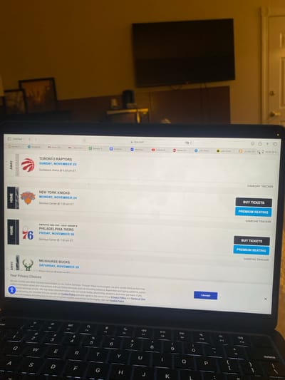 This image, taken in the City of Albany, United States, captures a close-up, angled view of an open dark-colored laptop displaying the NBA.com website, with a dimly lit interior room visible in the background. The laptop screen shows a list of upcoming NBA games, including "TORONTO RAPTORS" on Sunday, November 23, "NEW YORK KNICKS" on Monday, November 24 at Barclays Center, "PHILADELPHIA 76ERS" (Emirates NBA Cup) on Friday, November 28 at Barclays Center, and "MILWAUKEE BUCKS" on Saturday, November 29. "BUY TICKETS" and "PREMIUM SEATING" buttons are visible next to the Knicks and 76ers listings. A "Your Privacy Choices" cookie consent banner is displayed at the bottom of the screen with an "I Accept" button. The laptop's illuminated keyboard is partially visible at the bottom of the frame. In the background, a large, dark flat-screen television is mounted on a light-colored wall. Below the television, a dark brown furniture piece holds an indistinct object. To the left, two framed pictures hang on the wall, and to the right, a light-colored door is partially open. No individuals are visible in the scene.