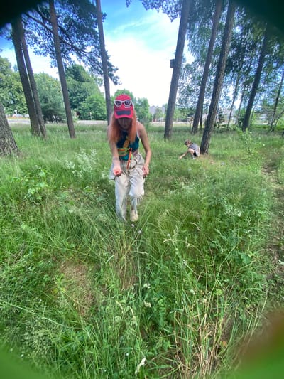The image depicts an outdoor scene with two individuals in a grassy area with scattered trees. In the foreground, a person with orange hair, wearing a red cap with sunglasses perched on top, a colorful top, and light-colored pants, is walking forward and holding a small red object in their hand. To the right and further back in the scene, a second person, appearing to be a child, is crouched down, interacting with the grass. The environment is characterized by tall grass, trees, and a bright sky with clouds. The overall impression is that of a casual, outdoor excursion.