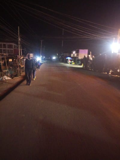 A dimly lit street scene at night features a man in a striped sweater walking towards the camera. Behind him, other figures walk further down the road. Streetlights create bright spots in the distance. Stalls line the left side of the road, while a large vehicle with a purple section sits on the right. Numerous power lines crisscross the sky above. The overall atmosphere is dark and blurry.