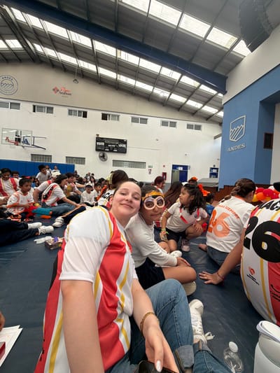 The image shows a group of people inside what appears to be a gymnasium or sports hall in Tlaxcalancingo, Mexico. A large group of people, including children, are sitting on the floor. Many individuals are wearing white shirts with red and yellow stripes. There is a girl who is holding the camera, smiling, who is wearing the same shirt. Some of the individuals have the number "6" on the back. A group of women in the background are wearing shirts with the number "6" and the phrase "Para Siempre" on them. The word "Andes" is visible on the blue wall. A digital scoreboard can be seen on the wall, as well as a basketball hoop. The gym has a high ceiling with skylights, providing a lot of natural light.