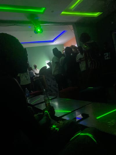 A dimly lit room features green and blue neon lights. Several people are seated, some wearing white clothing and head coverings. A clear bottle is visible in the foreground, illuminated by green lasers. A dark, indistinct object occupies much of the lower portion of the frame. The room's interior has a modern design with linear lighting elements.