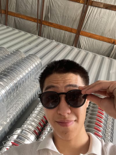 In Кенен Әзірбаев ауылы, Kazakhstan, a person with short dark hair and wearing sunglasses and a white polo shirt is taking a selfie. The individual is looking towards the camera, with their right hand adjusting the sunglasses. Behind the person are stacks of industrial metal ducts. The environment appears to be indoors, with a corrugated metal ceiling and wooden beams visible.
