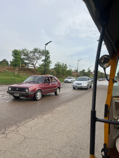 The image presents a street scene in Karmajiji, Nigeria, viewed from within a vehicle, likely an auto-rickshaw, as suggested by the yellow frame and side mirror in the upper left. The road is asphalt, appearing wet and possibly reflecting light, indicative of recent rain or damp conditions. The sky is overcast and cloudy.
Traffic is visible, with several cars on the road. A prominent maroon or red older model Volkswagen Golf is in the foreground on the right, displaying a Nigerian license plate "UYI-87JT". Further down the road, a silver Toyota Corolla is visible, along with other lighter-colored vehicles in a line. The road is flanked by green vegetation, including trees and grass, and streetlights with dark fixtures are spaced along the side. Electrical wires are also visible in the distance. The time of day appears to be daytime, but the heavy cloud cover prevents strong shadows. The scene depicts a typical road environment with vehicles in transit or stopped in traffic.