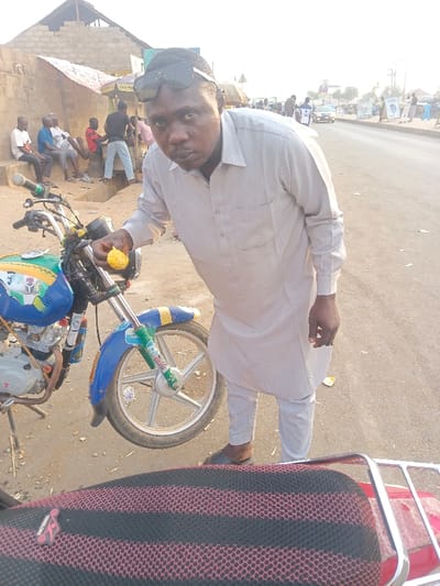This full-shot image captures a man standing beside a motorcycle on a street in Alizaga, Nigeria, during daytime. The man, with sunglasses pushed onto his head, wears a light beige or grey long-sleeved tunic (likely a traditional kaftan or kurta) and matching trousers, paired with dark slippers. He is looking directly at the camera with a serious expression, holding a yellow, textured food item in his right hand.
To his left is a vibrantly decorated motorcycle, predominantly blue and green, with numerous stickers and custom elements on its tank, front fender, and suspension forks. The engine casing has a "TVS" logo visible, and the front wheel features silver multi-spoke rims. In the immediate foreground, the red and black hexagonal mesh seat of another motorcycle is partially visible, with "TEMPLONG" text on a small tag.
In the background, several men are casually sitting and standing near simple block-wall buildings with corrugated iron roofs, indicative of a roadside or market setting. The road surface transitions from dirt or unpaved shoulder to asphalt further in the distance, where more people and vehicles are faintly visible. The natural lighting suggests a clear day, possibly late morning or early afternoon. The scene portrays a typical bustling street environment.