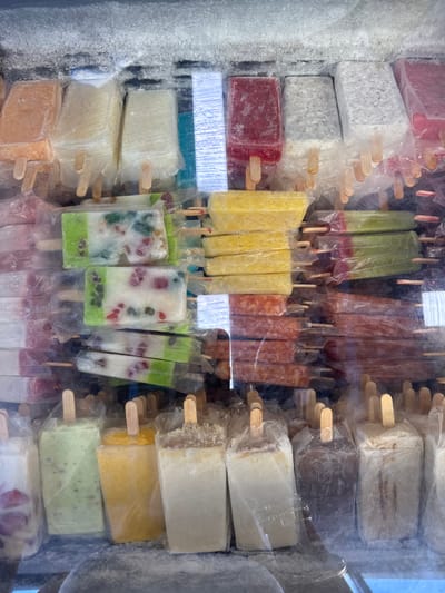 The image shows a refrigerated display case with a variety of popsicles, in a small grocery store or street vendor stand. There are numerous rows of different flavors and colors, including red, white, blue, yellow, green, and brown. The popsicles are arranged neatly in plastic wrap with wooden sticks protruding from the top. The display case is located in a small shop in Ciudad Santa Catarina, Mexico, at coordinates 25.658 latitude, -100.439 longitude. The interior of the shop is not visible. The condensation on the glass case suggests that the temperature is cool, which would be expected for a refrigerated space. The scene depicts a typical food vendor in Mexico, likely offering popsicles as a popular, refreshing treat.