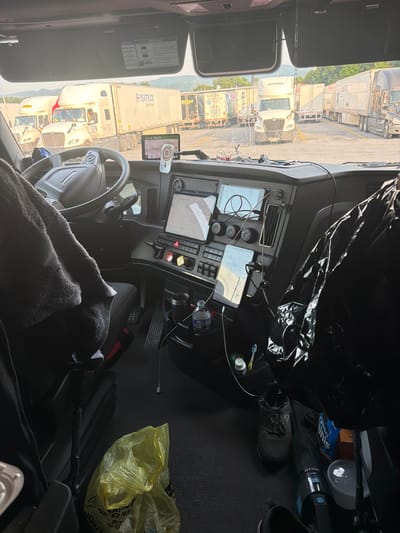 The image shows the interior of a semi-truck cab, parked outdoors. The driver's seat and dashboard are visible, along with various personal items and truck accessories. Outside the cab, several other semi-trucks and trailers are parked in a lot, with some vegetation and hills in the background. The scene suggests a rest stop or truck yard. The sun appears to be shining, indicating daytime. The specific location is identified as Raphine, United States.