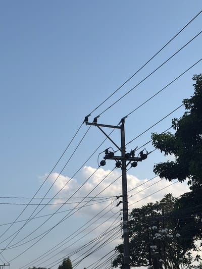 The image captures a view of a utility pole with multiple electrical wires crisscrossing against a bright blue sky with scattered white clouds. The pole is made of wood and supports several ceramic insulators, which hold the wires in place. To the right, a lush green tree with some white flowers is partially visible, its branches reaching towards the wires. The scene appears to be outdoors in a location that could be found within the city of Magelang, Indonesia, given the tropical foliage and clear sky. No people or vehicles are visible in the frame. The overall impression is one of an everyday urban or semi-urban landscape, focusing on the infrastructure of electrical distribution and the surrounding natural elements.