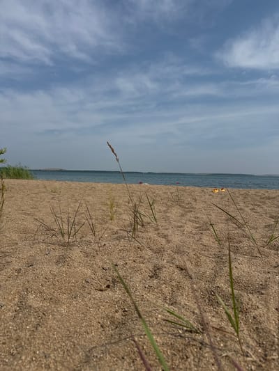 The image depicts a close-up view of a sandy beach with sparse dry grass in the foreground. The sand is light brown and appears somewhat coarse. In the background, a calm body of water stretches to the horizon, with a hazy line of land visible in the distance. The sky above is a pale blue with scattered white, wispy clouds. A few red buoys are visible in the water. On the sand, near the water's edge, a pair of yellow flip-flops is visible. The scene suggests a tranquil, possibly deserted, beach location. The mention of Ayyrtau, Kazakhstan, places this scene within a specific geographical context, likely a natural lake or riverfront within that region.