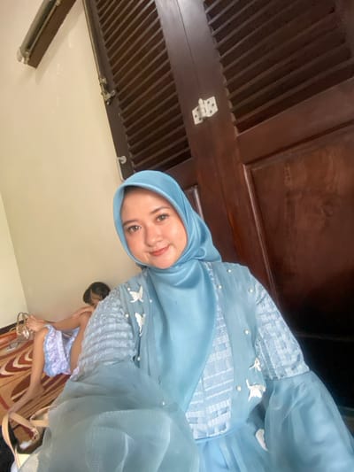 An indoor photograph, taken in the city of Ketegan, Indonesia, features a female individual positioned in the foreground, facing the camera with a faint smile. She is wearing a light blue hijab and a matching light blue formal dress or top, which includes textured fabric on the sleeves and bodice, adorned with white pearl-like beads and white butterfly appliques. The sleeves are voluminous and constructed from sheer netting. In the mid-ground to the left, a second individual, partially obscured and possibly a child, is lying or sitting on a patterned rug, dressed in a light-colored garment. A light-colored handbag with a gold-colored chain handle rests near this individual. The background on the right side of the image consists of a dark brown wooden door featuring vertical slats and visible white metal hinges. The wall in the background to the left is a plain light color.