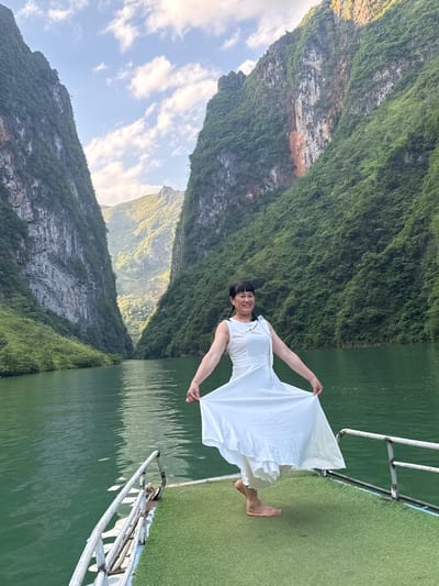 A woman in a white dress stands barefoot on a green, artificial-turfed platform with a white railing, holding the edges of her dress and smiling. The platform appears to be the deck of a boat on a river or lake, as water surrounds it. Towering, tree-covered mountains rise steeply on both sides of the water. The sky is partly cloudy, with sunlight illuminating the landscape. This scene is likely located in Đồng Văn Commune, Vietnam, known for its dramatic karst landscapes.