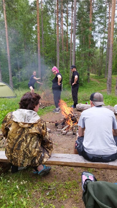 In a forest clearing, a group of four individuals gathers around a crackling campfire. The scene is set in a wooded area with tall, slender pine trees and lush green foliage. A green tent is visible in the background to the left.
Two people are seated on a rustic wooden bench, facing away from the camera, towards the fire and the other two individuals. One person, wearing a camouflage jacket, is seated on the left, while the other, wearing a grey t-shirt and a baseball cap, is on the right.
Standing near the campfire are two other individuals. One person, wearing a black t-shirt and black pants, has their hands on their hips. To their left, a person with vibrant pink hair, wearing a black t-shirt and black pants, stands with their hand on their hip. A fourth person, also wearing a dark t-shirt and shorts, is tending to the fire, reaching towards it with a branch. Smoke billows from the fire, partially obscuring the background.
The lighting suggests it is daytime, likely afternoon, with overcast or diffused light filtering through the trees. The overall atmosphere appears relaxed, with people enjoying a moment in nature. The location cues of tall pines and dense undergrowth are characteristic of a Russian forest, fitting with the provided context of Orekhovo, Russia. There are no visible signs or text in the image.