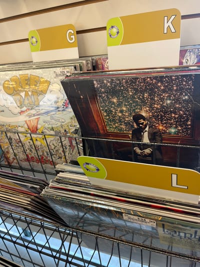 This image captures a retail display of vinyl records inside a store, presumed to be located in Edmond, United States. The records are organized vertically within black wire rack bins, segmented by large, yellow and white alphabetical divider cards marked 'G', 'K', and 'L'. Each divider card features a circular green and yellow 'BUY SELL TRADE' logo. Prominent albums include 'Green Day - Dookie' on the left, displaying an elaborate cartoon cover and a $29.99 price tag. Adjacent to it is 'Kid Cudi - Man on the Moon II: The Legend of Mr. Rager', depicting a seated figure within a wooden frame against a dense starry background. A partial view of 'Lamb of God - Ashes of the Wake' with a $34.99 price tag is visible in the lower right bin. The background consists of a light-toned wall with horizontal paneling.