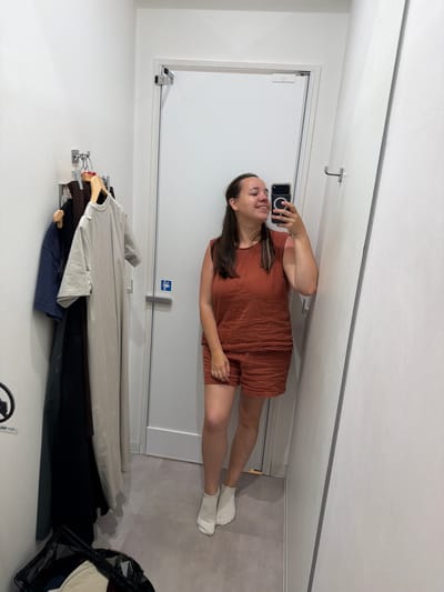 In a fitting room in Chuo, Japan, a woman stands and takes a selfie in a mirror. She is wearing a rust-colored linen-blend top and matching shorts. Her hair is dark brown and pulled back. To her left, several garments hang on a rack, including a long, light-colored dress, a dark blue t-shirt, and a black garment. A black mesh laundry basket sits on the floor in the lower left corner of the image. The walls of the fitting room are white, and the floor is tiled.