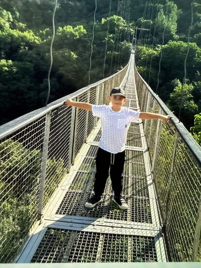 A young boy wearing a white t-shirt with a subtle pattern, black pants, a baseball cap, and sneakers stands on a suspension bridge. He is facing the camera with his arms outstretched, leaning slightly on the railing. The bridge has a metal grating walkway and railings made of metal mesh. It extends into the distance, surrounded by lush green trees and foliage, suggesting a natural, possibly mountainous environment. The lighting indicates it is daytime, with shadows cast on the bridge. The background features dense forestation and the continuation of the bridge structure disappearing into the trees.