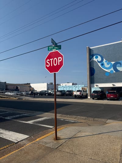 This media file depicts an outdoor scene with a prominent stop sign in the foreground. The stop sign is red and octagonal with white lettering spelling "STOP." Attached to the same pole as the stop sign are two street signs. One sign is green and reads "WAYNE," and the other is also green and reads "7TH STREET."
The scene is set on a street with a sidewalk and a parking area. Several cars are parked along the side of the street, including a red SUV, a black SUV, and a white pickup truck. The background features buildings with a blue mural on one of them, adorned with a swirling white and blue wave design. The sky is clear and bright blue, suggesting a sunny day. The lighting indicates it is likely daytime, possibly late morning or afternoon.
The image appears to capture a typical urban street environment. There is no obvious activity taking place beyond the presence of parked cars and the static street signs. The overall impression is one of a bright, clear day in a commercial or residential area. No people are clearly visible or identifiable as the primary subject of the image. The focus is on the street signage and the immediate surroundings.
