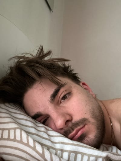 A close-up, angled shot depicts a Caucasian adult male subject situated indoors in Rafz, Switzerland. The man, captured from the chest up, is lying on his left side with his head resting on a white pillow featuring beige horizontal stripes. He has medium-brown, disheveled hair, a beard, and a mustache. His right eye is open, while his left eye is partially closed. His lips are slightly parted. A thin, gold-toned chain is visible around his neck on the right side of the frame. The background consists of a plain, light-colored wall, with a partial reflection of an unseen object or frame visible in the upper left quadrant. No other individuals or notable objects are present. The ambient lighting is even.