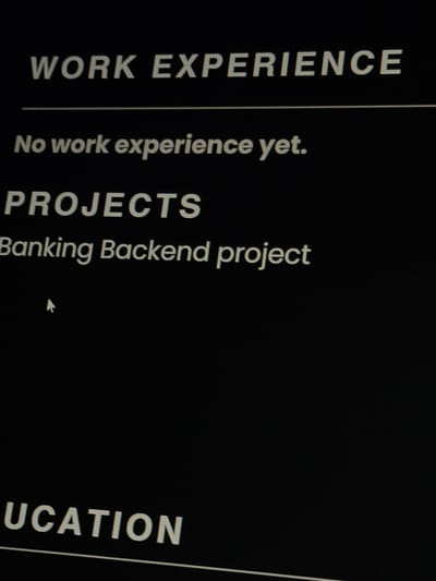 The image displays a dark screen, likely a digital display or a printed document on a dark background, featuring sections of a resume or curriculum vitae. The overall tone is monochromatic, with light gray/white text against a black background. The image is slightly rotated.

Visible text includes headings and content sections:
- A prominent heading reads "WORK EXPERIENCE", presented vertically along the left edge.
- Below this section, the text "No work experience yet." is displayed.
- Another heading, "PROJECTS", is visible further down.
- Under "PROJECTS", the line "Banking Backend project" is present.
- Towards the bottom right, a partial heading "UCATION" is seen, strongly suggesting it is part of the word "EDUCATION".

A thin vertical line acts as a visual separator between the main content area and what appears to be a sidebar or another section. A white mouse cursor, shaped like an arrow, is visible pointing towards the text "Banking Backend project". The image appears to be a photograph of a screen, indicated by the slight glare and digital noise. The setting is indoors, with no specific time of day or weather cues discernible from the image content itself, beyond the dark screen implying a potentially low-light environment or a design choice. The photo was taken in Mkhchyan, Armenia.