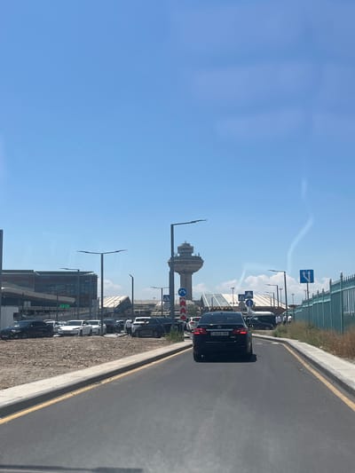 The image depicts an outdoor scene on a clear, sunny day, likely late morning or early afternoon, given the bright sunlight casting shadows. The setting appears to be an airport or a similar transportation hub, with a control tower visible in the background. A road with a yellow line marking the edge runs along the right side of the frame, bordered by a gravel and concrete area. A row of parked cars is visible along the left side, some of which are white and one black car with a license plate that reads "16 EE 999". Several traffic and directional signs are also present, including one with arrows indicating a left turn and going straight, and others with symbols that are not clearly decipherable. In the distance, a tall, modern building complex can be seen, along with a fence and some dry vegetation. The overall atmosphere is one of everyday activity at a transportation facility. There are no people clearly visible in the image, though some figures in white attire might be airport personnel in the distance.