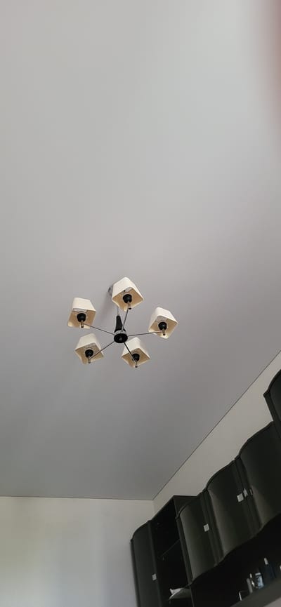 The image shows a modern five-light chandelier with cream-colored lampshades hanging from a light gray ceiling. The chandelier has a central black hub with thin metal arms extending outwards, each holding a lampshade. The lampshades are square with an open bottom, revealing the light bulb sockets. The chandelier is positioned slightly off-center in the upper portion of the frame. Below the ceiling, a white wall is visible, transitioning to a dark, curved cabinet or shelving unit that occupies the lower right portion of the image. The cabinet appears to have a glossy finish and contains some items on its shelves, though they are not clearly discernible. The image was taken in an indoor environment, likely a room. The location of Bulgakovo, Russia is not determinable from the visual information presented in the image.