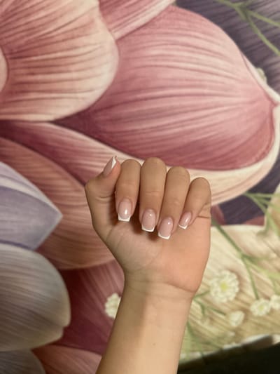 A human left hand is positioned centrally, oriented with the palm slightly upward and fingers gently curled, displaying a freshly applied French manicure. Each fingernail features a translucent pink-beige base with a distinct white tip. The skin tone of the hand is light to medium. The background consists of a diffused, multi-toned floral pattern, predominantly featuring large, blurred petals in shades of pink, purple, and cream, characteristic of wallpaper or a tapestry in an indoor setting. The location is Dmitriyevskaya, Russia.