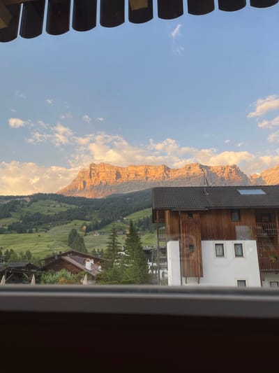 The image depicts a scenic mountain landscape viewed from a building. The primary subjects are the dramatic, sun-drenched mountain range and a charming alpine village nestled in a lush green valley. The mountain faces are illuminated by the warm, golden light of what appears to be either sunrise or sunset, highlighting their rugged textures and dramatic cliffs.
The village features traditional wooden chalets and houses, interspersed with green meadows dotted with smaller structures, likely farmhouses or guesthouses. Dense green forests climb the slopes of the mountains, creating a striking contrast with the rocky peaks. The sky above is a clear, pale blue, with scattered white clouds that are also catching the warm light.
The perspective suggests the photo was taken from a window or balcony of a building. A portion of the building's structure, including wooden paneling and windows, is visible in the foreground, framing the view. On the left side of the image, there are dark, curved objects that appear to be part of an external shading system or a balcony railing, creating a sense of looking out from an interior space.
The overall mood is serene and idyllic, evoking a sense of peacefulness and the beauty of nature. The time of day is clearly indicated by the low angle of the sun casting long shadows and bathing the mountains in a warm glow. The location, identified as La Ila - Stern - La Villa, Italy, aligns with the visual cues of alpine architecture and dramatic mountainous terrain. No text is visible in the image.