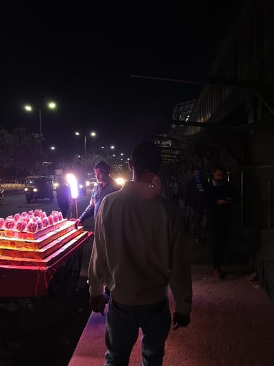 The image depicts a street scene in Noida, India, at night. The location is near the Sector 18 Metro Station (latitude: 28.587, longitude: 77.375). A street vendor, wearing a blue shirt, is standing beside a red cart containing boxes of what appears to be fruit. The vendor is looking at the camera. A man, wearing a light brown jacket, is facing away from the camera. His back is to the viewer, he is looking at the fruit vendor, and his hand is outstretched toward the cart. Three people are visible in the background walking past, two of them are blurry. There are streetlights casting light on the street. There is a dark building to the right of the scene. The sidewalk is lit by the streetlights, and the ground is a mix of concrete and asphalt. There is a car in the background, and the environment is dark and dimly lit. The image is a night scene taken at an urban street location.