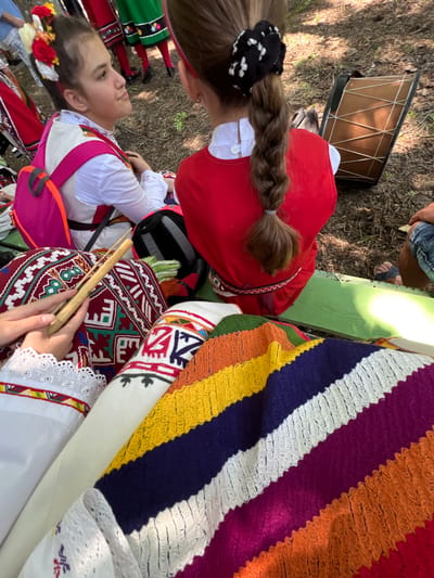 The image depicts an outdoor cultural gathering in Bolyarovo, Bulgaria. Two young females, dressed in traditional Bulgarian folk costumes, are prominent in the foreground and midground.
The girl on the left, viewed in profile, wears a white embroidered blouse with lace trim, a light-colored vest, and has her dark hair pulled back and adorned with red, yellow, and white fabric flowers. A bright pink backpack with orange straps is worn over her costume. She is seated, looking towards the right with a serene expression.
The girl on the right, viewed from behind, has her brown hair styled in a long braid, secured at the top with a black and white polka-dotted scrunchie and at the end with a grey elastic band. She wears a red sleeveless garment or pinafore over a white collared shirt, with a patterned belt featuring geometric designs around her waist. She is seated on a green wooden bench, part of which is visible beneath her.
In the bottom left foreground, a pair of hands with a white, lace-trimmed sleeve holds a light wooden stick-like instrument, possibly a shepherd's flute (kaval) or similar, which rests on a richly embroidered red textile featuring geometric patterns in white, green, and blue. Adjacent to this is a folded, multi-colored striped textile, likely a blanket or shawl, displaying bands of yellow, navy blue, white, magenta, orange, and red.
Behind the girl on the right, a traditional wooden frame drum (likely a davul or tapan) with visible rope lacing is lying on the uneven, earthen ground. The ground itself is covered with dry leaves and sparse foliage, indicative of an outdoor, possibly wooded, setting. A partial view of a person's leg in blue jeans and brown sandals is visible to the right of the girl on the bench. In the blurred background, other individuals are partially visible, some also in traditional folk attire, including red and green skirts with white embroidery, suggesting a larger assembly or performance. The scene is illuminated by natural daylight.