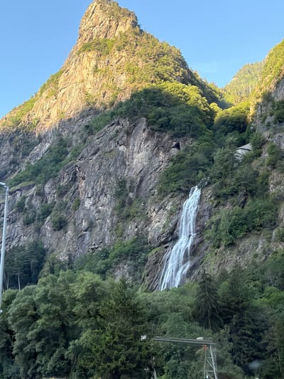 This image depicts a natural landscape near Miéville, Switzerland. A prominent, sheer rock face dominates the background, with a tall waterfall cascading down its rugged surface. The mountain is partially illuminated by sunlight, casting shadows across the textured rock. Lush green vegetation, including dense trees and shrubs, covers the lower slopes of the mountain and the foreground. The sky above is clear and blue. In the lower left of the image, a portion of a white lamppost is visible, suggesting proximity to a road or developed area. In the lower right, a section of a metallic structure, possibly part of a fence or an elevated walkway, is also present. The overall scene is one of natural grandeur with subtle hints of human infrastructure.