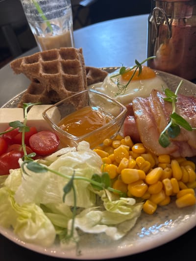 This media file shows a close-up, top-down view of a breakfast or brunch dish served on a patterned plate. The dish appears to be a savory and sweet combination. On the right side of the plate, there is a generous portion of bright yellow corn kernels. Adjacent to the corn is a pile of shredded iceberg lettuce, adding a fresh green element. In the center of the plate, there's a small, clear glass dish filled with a thick, amber-colored jam or preserve, possibly apricot or peach. To the left of the jam, there are halved red cherry tomatoes and small cubes of white cheese, likely feta or mozzarella. A piece of toasted rye waffle, with its distinctive grid pattern, is positioned on the bottom left of the plate. Above the jam and cheese, there's a fried egg with a slightly runny yolk, accompanied by slices of crispy, cooked bacon. Delicate green sprouts are sprinkled over several components of the dish, adding a garnish. A portion of a metal cup or container with a handle is visible at the top left of the frame, possibly indicating it's part of a beverage or another dish. The lighting suggests an indoor setting, likely a restaurant or cafe. The overall presentation is appealing, highlighting a variety of textures and flavors. There is no visible text in the image. The time of day appears to be morning or midday, suitable for breakfast or brunch.