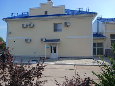 The image shows a light yellow, two-story building with a blue tiled roof under a clear blue sky. The building has multiple windows and air conditioning units mounted on its exterior walls. There is a white door with a ramp leading up to it. In the foreground, there are dark red leafy bushes. The building appears to be part of a complex, with another similar structure visible to the right. The ground in front of the building is paved with grey interlocking stones and concrete slabs. A blue sign with white text is visible on a pole in front of the building. The overall scene is one of a modern, well-maintained building in an outdoor setting, likely in a suburban or industrial area given the architecture and paving. The location is identified as Chazhemto, Russia.