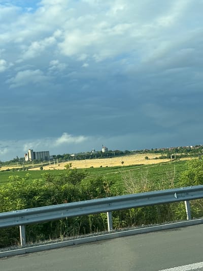 The image captures a rural landscape with a cloudy sky. In the middle ground, a large field of hay bales is visible, with rows of green crops in the foreground. In the distance, a cluster of buildings, possibly industrial silos and some houses, are situated on a gentle slope. A road barrier is present in the immediate foreground, suggesting the vantage point is from a road or highway. The overall scene depicts a cultivated agricultural area under a dramatic, overcast sky.